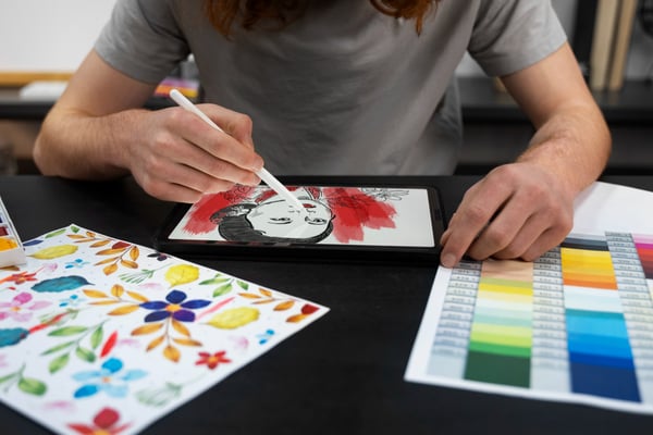 Photo of selling artwork as a side hustle idea shows a person sitting at a table, drawing on a tablet. On the table, there is a drawing of flowers and a color palette.
