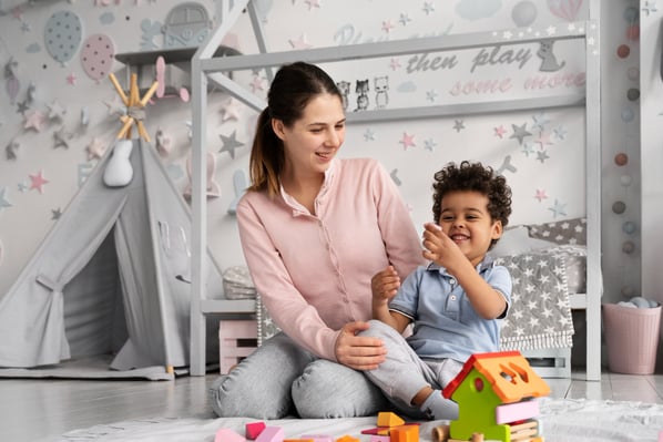 Photo that illustrates blog post with side hustle ideas shows a woman sitting on the floor with a toddler, playing with a wooden house in a kid's bedroom.