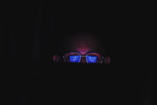 Picture shows a man in a dark room in front of a computer screen and wearing glasses. The blue light from the monitor is reflecting on his glasses' lenses, illustrating blue light-blocking glasses as some of the best dropshipping products.