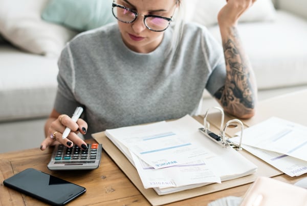 Photo shows a woman wearing glasses and with tattoos on her left arm sitting on the floor in a living room, looking at invoices and using the calculator on a coffee table. This picture is part of Shoplazza's article on what is break-even analysis.