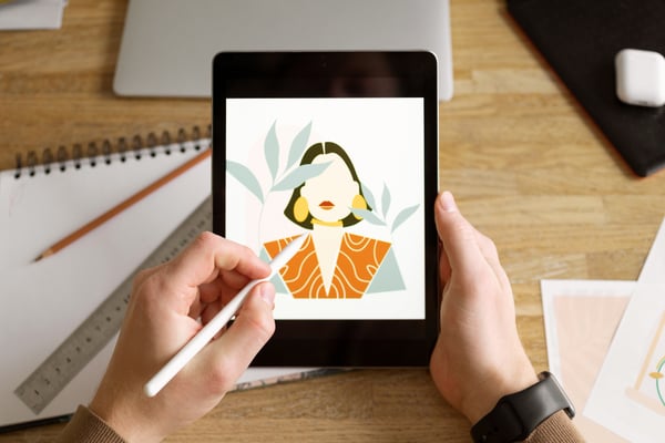 Photo from article about selling digital products online shows a person holding a tablet and a tablet pen. The person is drawing an illustration of a woman with the tablet.  