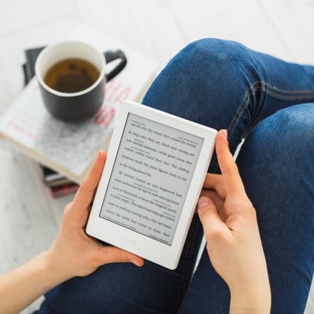 Picture shows a person sitting down, holding an ereader and reading an ebook. There is a pile of physical books on the floor next to them, and there's a cup of tea on top of the pile. This photo illustrates the article on how to sell digital products online.