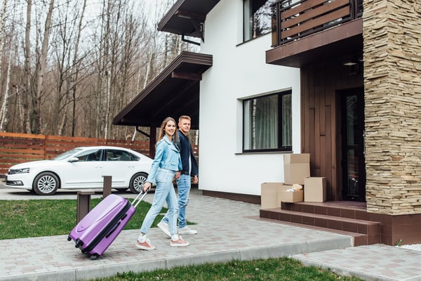 A couple holding hands, carrying a purple suitcase while entering a house, representing a short-term rent as a way you can earn money from home.