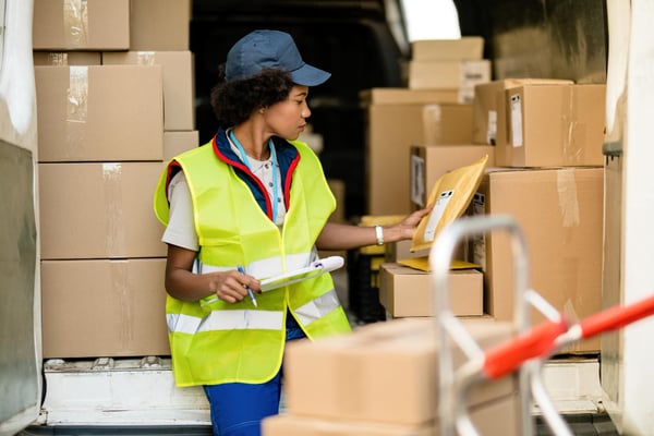 Photo shows a black person in a safety vest, holding a notepad and a packet. The person is surrounded by cardboard boxes. Finding suppliers is an important step in the process of how to start a dropshipping business.