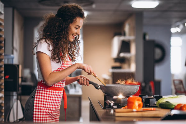 Photo shows a woman with long curly hair standing in front of a stove in a home kitchen, stirring food in a wok. Creating a food business is one of this article's side hustle ideas
