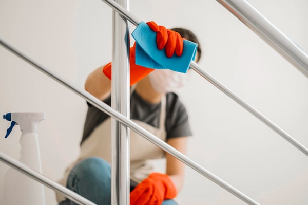 Photo shows a person wearing a face mask, rubber gloves and wiping a staircase handrail as an example of how house cleaning can be a side hustle idea to make extra money.