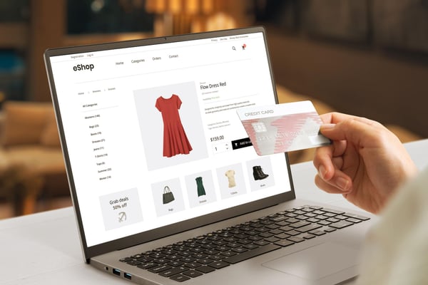 Picture illustrates article on how to start a dropshipping business. Photo shows a person sitting at a table, using a laptop to buy a red dress online. The person is holding a credit card.