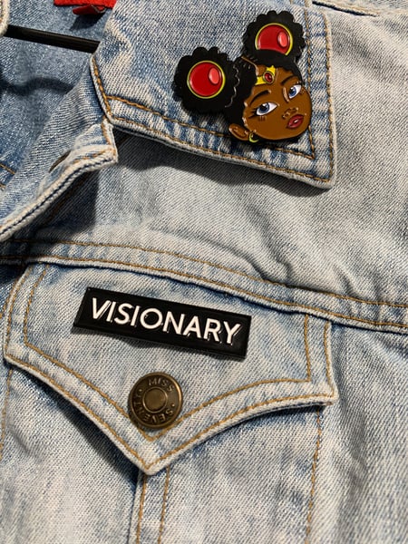 Picture illustrates what soft enamel pins look like, to help readers understand how to make an enamel pin. The picture shows a denim jacket from up close, with a black girl pin on the lapel and a pin that says visionary on the pocket.
