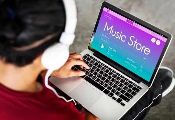 Photo taken from above shows a person with a laptop on their lap. The person has headphones on and they're looking at a Music Store page on the laptop. Picture is part of the How to sell digital products online in 6 steps section.