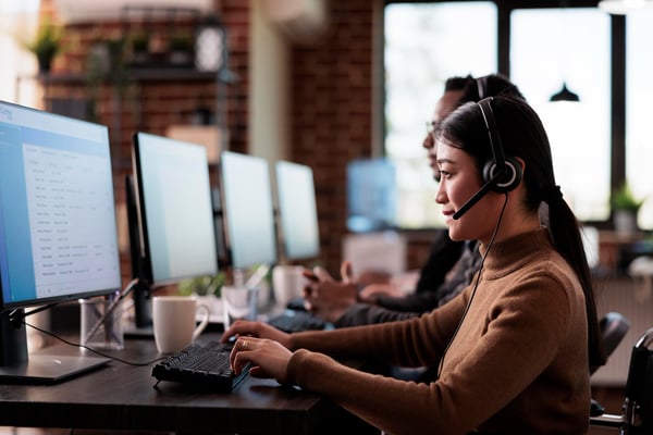 Photo shows people working in front of a computer, wearing headsets and smiling, to highlight the importance of improving customer service skills.