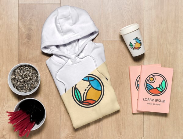 Photo shows a hoodie, a reusable coffee cup and 2 notebooks on a table with a mockup logo on them. Photo helps to show how you can make your own merch to sell online.