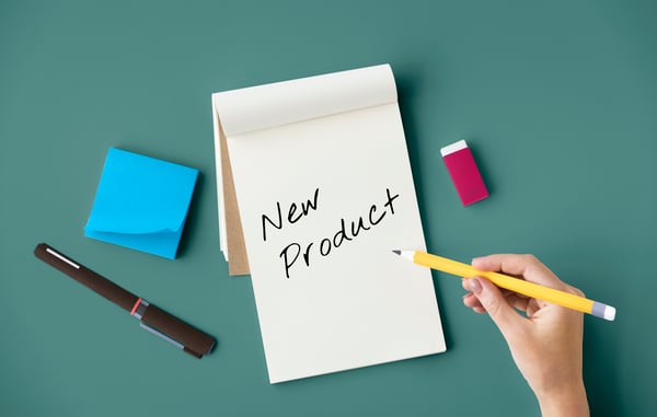 A notepad with the words "New Product" written in the middle, with a hand holding a pencil, a pen, a post-it and a eraser on a green table.