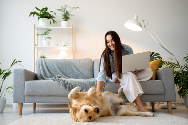 Photo that illustrates pet sitting as one of the side hustle ideas shows a woman with long hair sitting on a sofa, holding a laptop while she's petting a golden retriever that's lying on the floor.