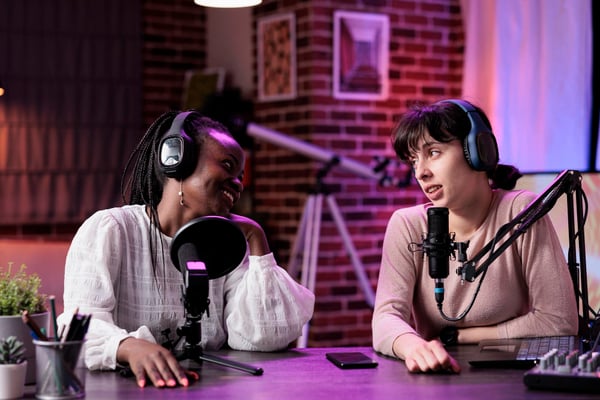 Photo shows two people sitting at a table, speaking into professional mics and wearing headphones. This photo alludes to podcasts, which is a way companies can invest in content marketing.