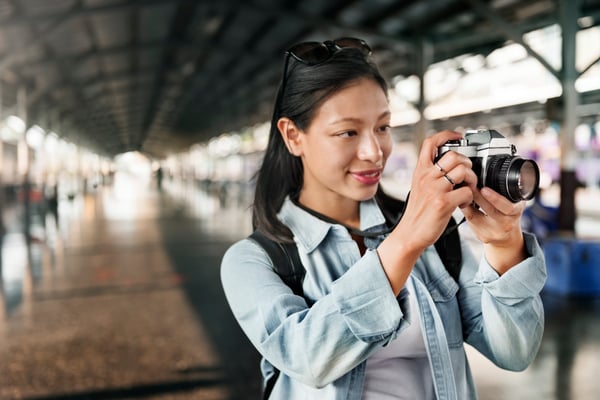Picture shows a woman with black hair at a public place, holding a professional camera. The photo alludes to the importance of taking professional product photos to sell enamel pins online.