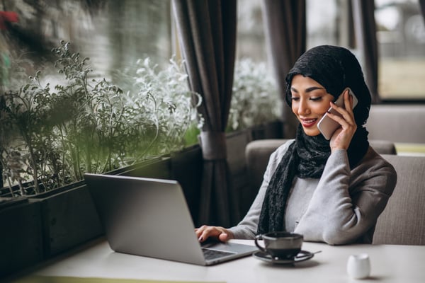 Photo shows a woman wearing a hijab sitting in a cafe, working on her laptop while using her phone. This photo illustrates the section Promote your business online in the blog post on starting an online business in Canada.