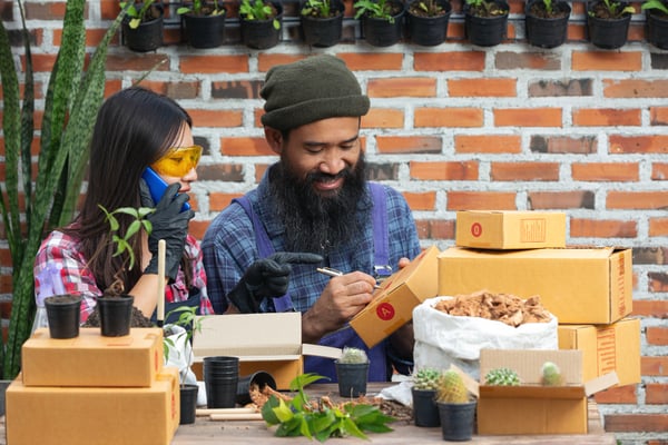 Photo shows two people working at a plant store. They are in front of a table covered with cardboard boxes and succulents. One of them is writing on the box, while the other is on the phone and wearing gloves. This photo illustrates article on selling plants online.