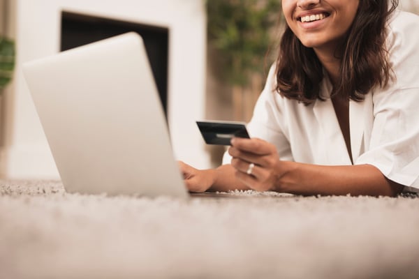 A woman using a laptop on the carpet, holding a credit card while smiling, representing that she's happy while shopping online
