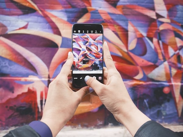 Photo illustrates blog post on starting an online business in Canada. In the photo, there is a person holding a phone and using the camera to take a picture of a colourful street mural.