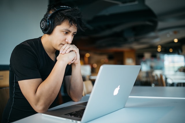 This photo shows a person sitting in a public place, possibly a cafe, in front of their laptop. They are wearing headphones and are staring at the screen. This photo illustrates section on how to start an online business in Canada by selling online courses.
