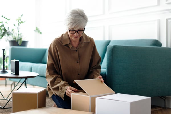 Picture shows a person in their sixties kneeling on the floor, surrounded by cardboard boxes. The person is lifting the lid of one of the boxes. This picture illustrates the idea of selling subcription services as a way to sell digital products online.