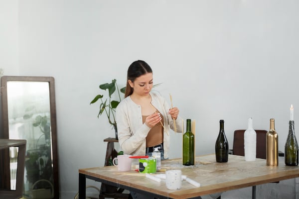 Photo illustrates article on green marketing. Photo shows a woman standing in front of a table, reusing glass bottles to make candle holders. She is gluing wooden sticks to the surface of the glass bottle to decorate it.