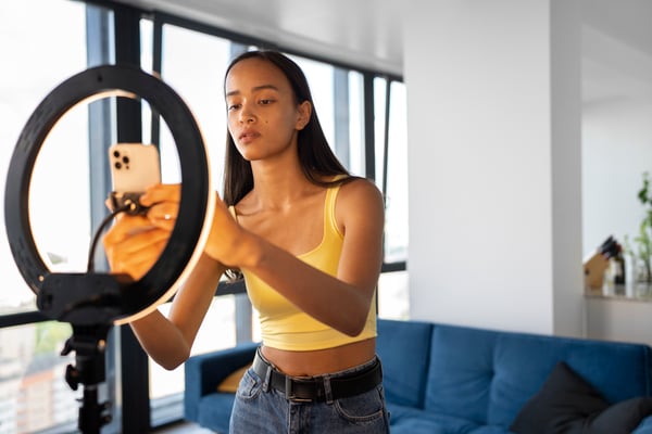 A brown-skin girl wearing a yellow t-shirt, conecting a celphone to a ring light, representing users creating their own TikTok Ads 