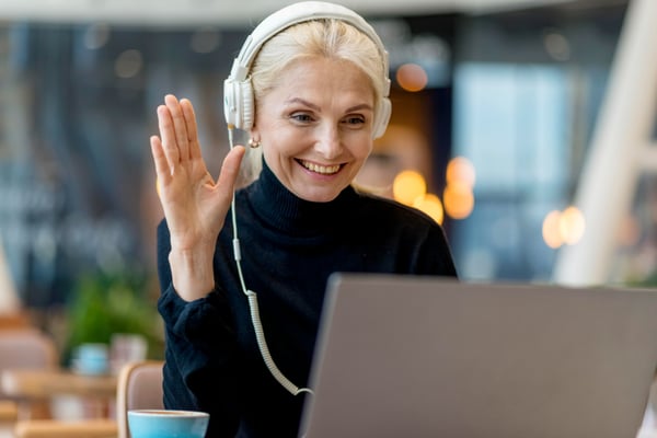 Photo in article about side hustle ideas shows a woman sitting at a table, waving at her laptop screen and wearing headphones.