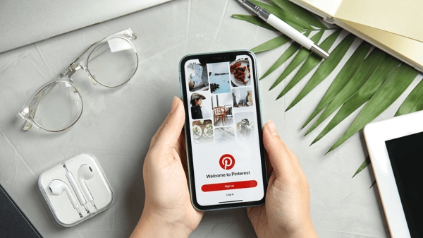 Picture illustrates article about how to make money on Pinterest. Picture shows a person's hands on a table-like surface, holding a smartphone. The phone screen shows the Pinterest log in page in the app.