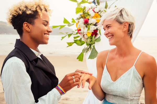 Picture shows two people getting married at the beach. One of them is sliding a wedding ring down the other's ring finger. Photo illustrates section of the article on picking a niche to start a jewelry business.