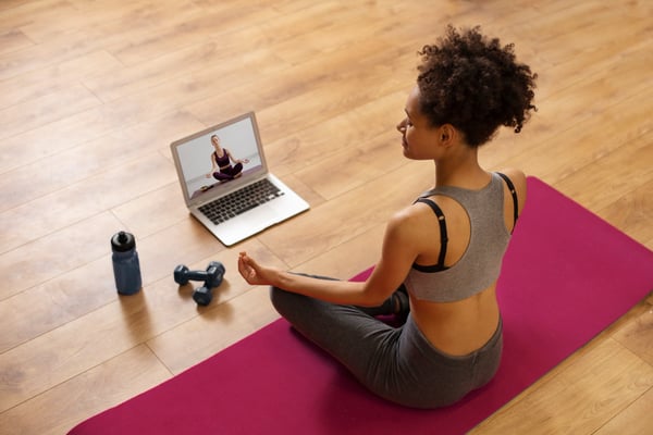 Photo illustrates article on digital products to sell online. Photo shows a person sitting on the floor, on a yoga mat, looking at a laptop screen that's also on the floor. The person is watching a yoga class, and both they and the instructor are doing the lotus position.