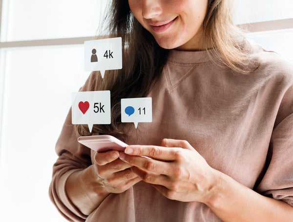 A blonde woman with white skin using a pink cell phone and social media icons of likes, comments and followers being displayed in boxes out of the cell phone screen, making reference to paid traffic that can be obtained through social media..