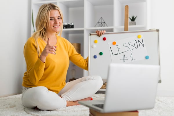 A blonde homan holding a board teaching a lesson online in a bright room while selling courses.